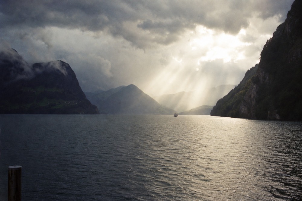 Raindrops on a boat window with blurred Lucerne lake shoreline and green hills beyond, no text, no logos, no people.