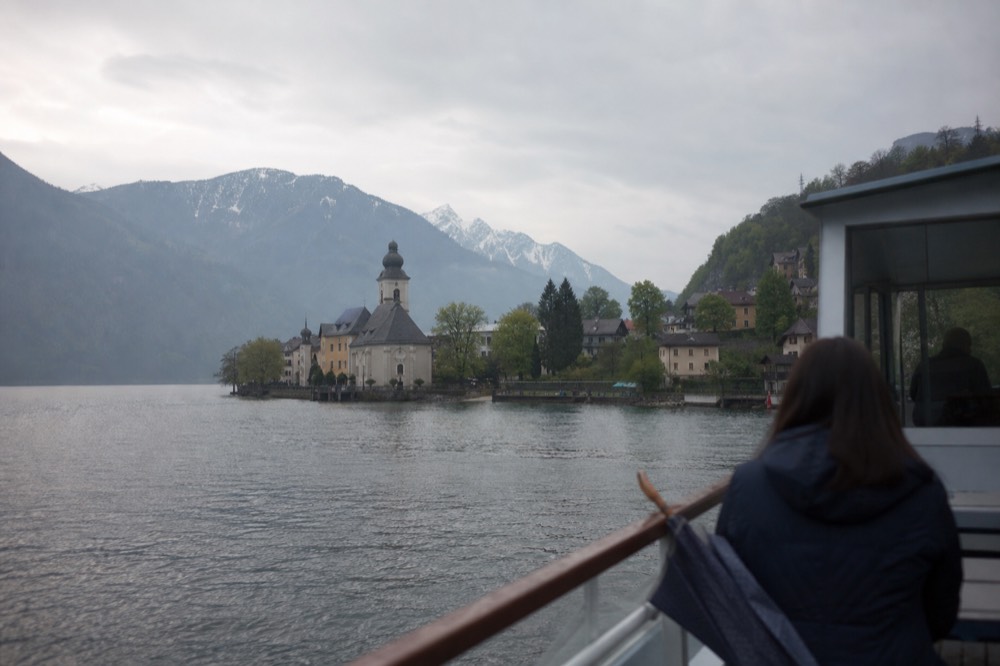 A cruise boat approaching a lakeside village with chapel and mountains, no text, no logos, no people
