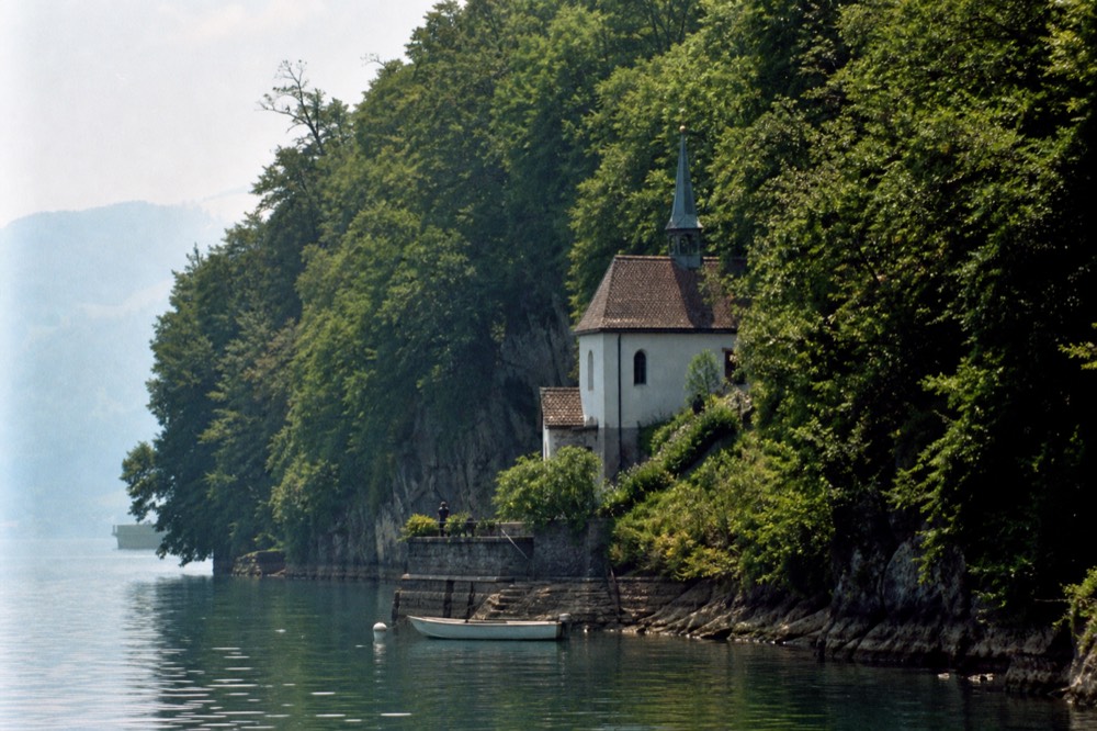 Steep forested shoreline of the Urnersee arm with a small white chapel above the water, no text, no logos, no people